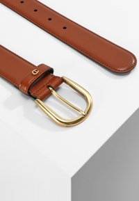 Bruine leren riem met een gouden metalen gesp, kenmerkend door een lichte textuur en verschillende gaten voor aanpassing. Versierd met een logo-detail.