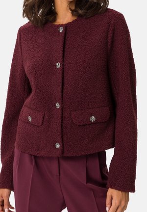 Kobieta w bordowej teksturowanej kurtce z srebrnymi guzikami w kształcie kwiatów oraz bordowych spodniach do kompletu.