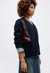 Γυναίκα με σγουρά μαλλιά που φοράει μπλε πουλόβερ με λαιμόκοψη σε σχήμα V πάνω από ριγέ πουκάμισο, φούστα από τζιν και κρατάει μπορντό δερμάτινη τσάντα ώμου.