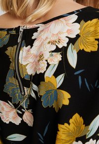 Blusa floreale con sfondo nero, decorata con fiori gialli e rosa. Ha un dettaglio di zip visibile in argento sul retro del collo.