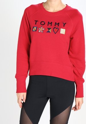 Personne portant un sweat-shirt rouge avec "TOMMY" et cinq patchs colorés, associé à des leggings noirs avec des panneaux en mesh transparent.