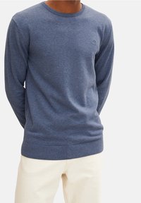 Blå stickad tröja med rund halsringning, långa ärmar och mudd i ärmsluten och nederkant. Subtil logotypdetalj på bröstet. Bärs med ljusa byxor.