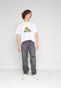 Mladý muž s kudrnatými vlasy, který má na sobě bílé tenisky, volné šedé roztrhané džíny a bílé tričko s grafikou šneka a nápisem "Take It Slow", na šedém pozadí.