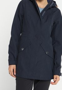 Granatowa wodoodporna kurtka z zamkiem błyskawicznym i zapięciem na guziki z przodu, z dwiema bocznymi kieszeniami i kapturem. Gładka tekstura z pionowymi przeszyciami.