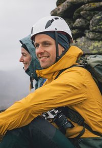 Gelbe wasserdichte Jacke, grauer Rucksack, weißer Helm. Zwei Personen sitzen, eine davon mit einer Kamera. Felsige Kulisse, bewölkter Himmel.