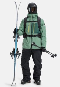 Skiausrüstung mit einer grünen Jacke mit gelben Reißverschlüssen, schwarzen Hosen, Helm und Skistöcken, die Skier mit blauen Streifen und Bindungen halten.