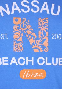 Niebieska koszulka z białym napisem "NASSAU BEACH CLUB" oraz pomarańczową dekoracyjną literą "N" z misternymi wzorami morskimi, data 2006 poniżej.