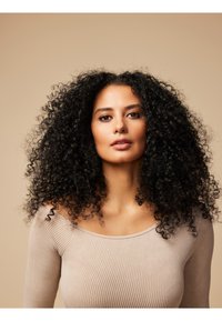 Portræt af en kvinde med fyldig sort krøllet hår, iført en lys beige strikket langærmet bluse, mod en neutral baggrund.