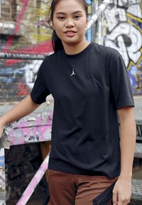T-shirt noir à manches courtes avec un petit logo blanc sur la poitrine, associé à un pantalon cargo marron, se tenant devant un fond de graffiti.