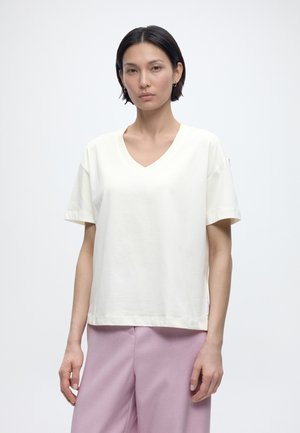 Giovane donna con capelli neri corti che indossa una maglietta bianca a V a maniche corte semplice e pantaloni lilla chiaro, in piedi davanti a uno sfondo bianco.