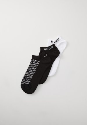 Trois paires de chaussettes de cheville : une noire avec le logo "HUGO" répété, une noire avec un seul logo, et une blanche avec le logo "HUGO" en noir.