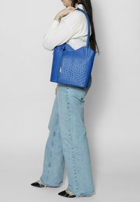 Sac fourre-tout en cuir bleu avec un motif texturé à pois, doté d'une fermeture éclair et de deux poignées. Porté à l'épaule avec un jean en denim.