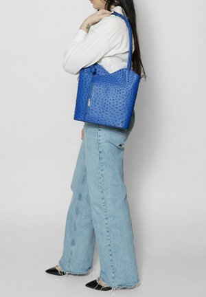Blauwe leren tote bag met een gestructureerd stippenpatroon, met een ritsluiting en dubbele handvatten. Gemaakt om over de schouder te dragen met denim jeans.