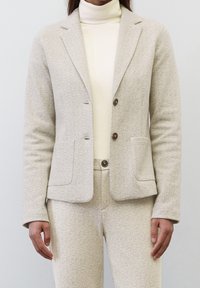 Světlý béžový blazer v rybí kostce s dvěma knoflíky, klopami, dvěma předními kapsami a přiléhavým střihem, nošený přes krémový rolák.