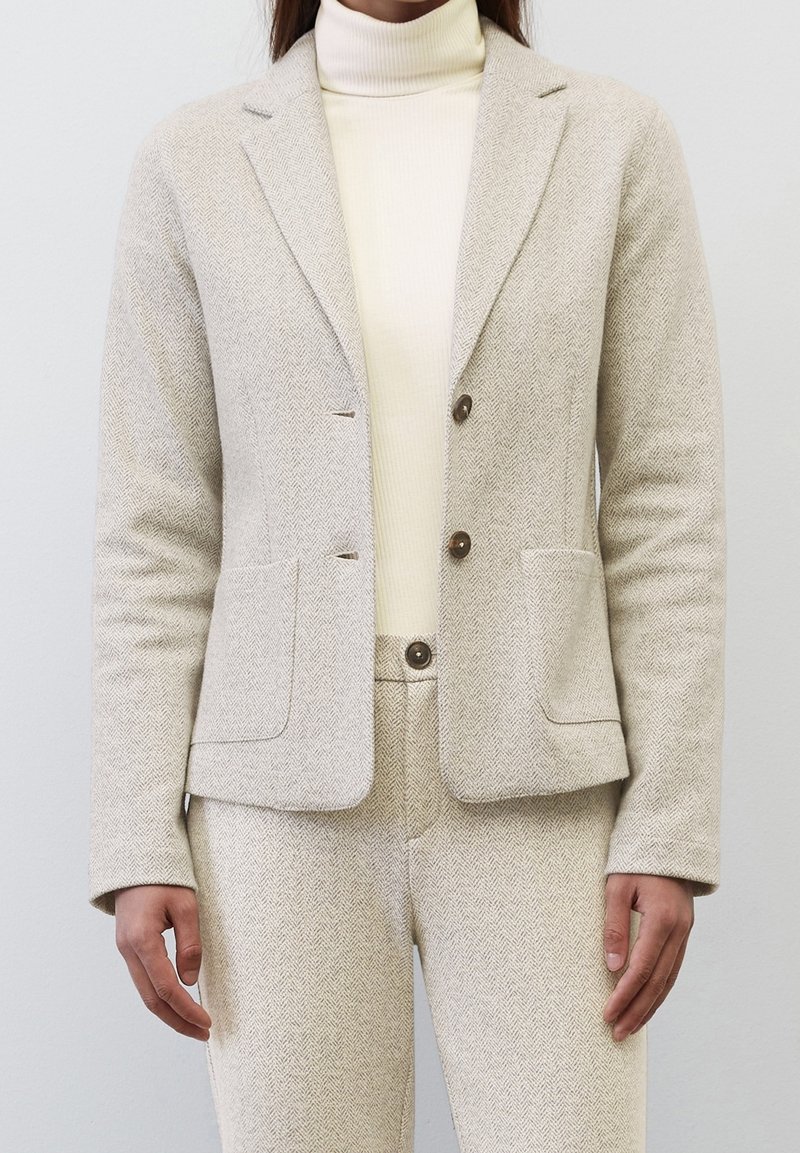Světlý béžový blazer v rybí kostce s dvěma knoflíky, klopami, dvěma předními kapsami a přiléhavým střihem, nošený přes krémový rolák.