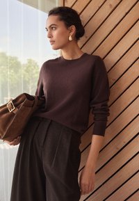 Brown knit sweater with a round neckline, paired with dark tailored trousers and a brown bag featuring a gold buckle, against wooden backdrop.