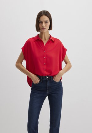 Woman with short brown hair wearing a loose red short-sleeve button-up blouse and dark blue jeans, standing with hands in pockets against white wall.