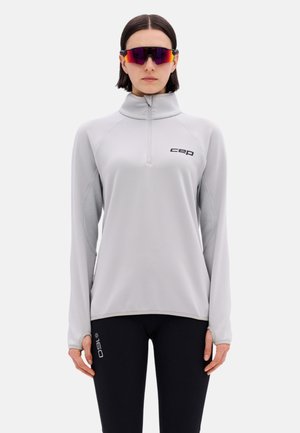Frau trägt hellgraues langärmeliges Zip-Up-Oberteil mit „cep“-Logo, schwarze Leggings mit der Aufschrift „160“ und reflektierende Sportsonnenbrille.