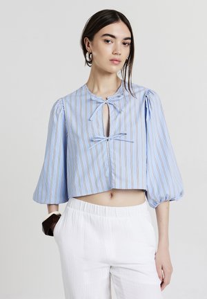 Junge Frau in einer blauen gestreiften, taillierten Bluse mit Puffärmeln und weißen hochgeschnittenen Hosen, die mit der Hand in der Tasche posiert.