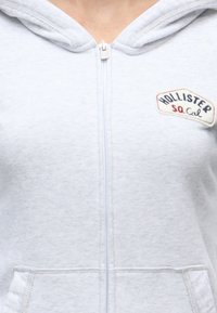White zip-up hoodie with front pockets and a Hollister So Cal logo patch on the left chest.