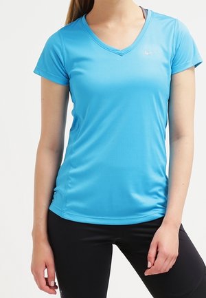 Vrouw draagt een lichtblauw sportshirt met korte mouwen en V-hals en zwarte leggings, staand met één hand ontspannen langs haar zijde.