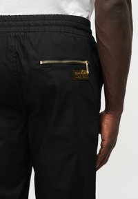 Inimene, kellel on mustad vöökohaga elastse vöökohaga püksid, lähedalt vaadatuna. Pükstel on kuldse lukk-taskuga silt "Versace Jeans Couture" taga.