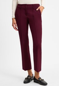 Pantalon bordeaux sur mesure avec une coupe slim, devant plat et deux poches latérales. Tissu lisse avec un léger éclat. Associé à des mocassins noirs.