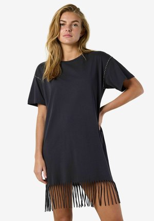 Femme aux longs cheveux blonds portant une robe T-shirt noire oversized avec des détails cloutés aux épaules et un ourlet à franges, posant avec une main sur la hanche.