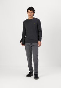 Suéter gris oscuro con cuello redondo y un detalle de logo amarillo, combinado con jeans grises y zapatillas negras, llevado con una bolsa negra.