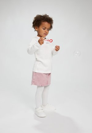 Weißes Langarmhemd mit dem Text "smile", rosa Jeansrock, weiße Strumpfhosen und weiße Sneaker. Kind, das mit einem roten Zauberstab Seifenblasen bläst.