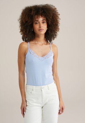 Un haut cami bleu clair avec un décolleté en V en dentelle. Fabriqué en tissu doux, il présente des bretelles fines et une coupe ajustée. Porté avec un pantalon blanc.
