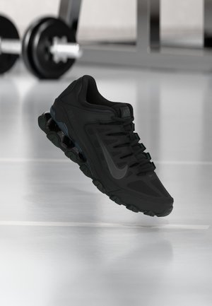 Zapatilla de running Nike negra con suela visible tipo resorte, colocada frente a un fondo de gimnasio con equipo de pesas desenfocado detrás.