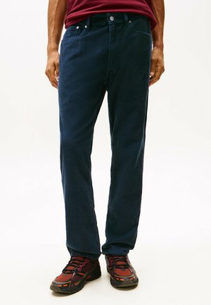 Marineblauwe corduroy broek met verticale ribbels, voorzakken en een knoopsluiting, gecombineerd met zwart-rode sportschoenen.