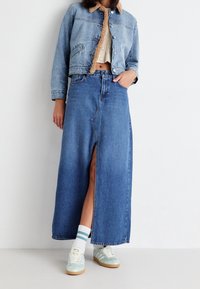 Jeansová bunda s hnědým límcem, rozepnutá vpředu, spárovaná s dlouhou jeansovou sukní s předním rozparkem. Světle modré tenisky a bílé ponožky.