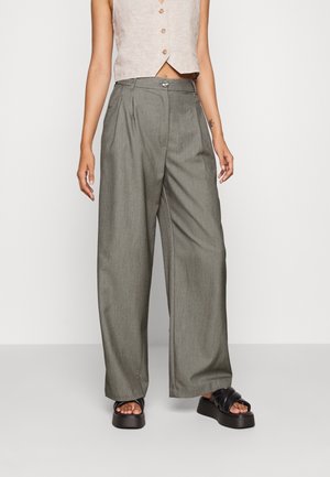 Persona que lleva pantalones grises de pierna ancha y cintura alta, top beige sin mangas con botones y sandalias negras de plataforma, de pie frente a un fondo liso.