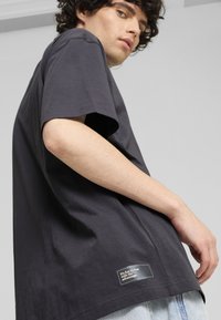 Camiseta de algodón gris oscuro con mangas cortas, que presenta una etiqueta discreta en el dobladillo con texto blanco y un diseño de corte relajado.