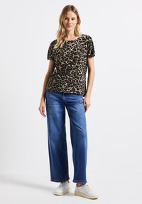 T-shirt con stampa leopardata in toni oliva, maniche corte, abbinata a jeans a gamba larga blu e sneakers bianche, su sfondo neutro.