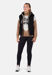 Gilet in pile nero sopra un maglione a collo alto beige con motivi geometrici, abbinato a jeans scuri e sneakers bianche.