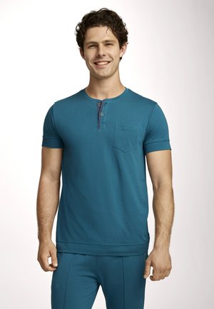 Lächelnder Mann mit lockigem Haar, der ein türkises kurzärmliges Henley-Shirt und passende Hose trägt und vor einem schlichten, hellen Hintergrund steht.