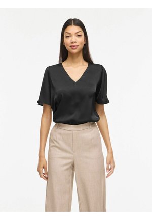 Kobieta ubrana w czarną satynową bluzkę z krótkim rękawem i beżowe, szerokie spodnie z wysokim stanem, stojąca na białym tle.
