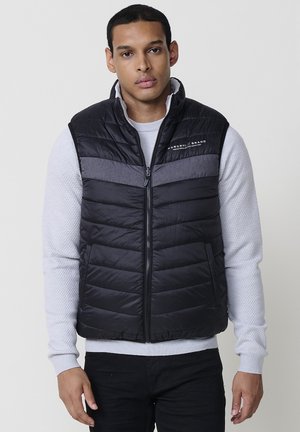 Chaleco acolchado negro con diseño de costuras horizontales, con una banda gris en el pecho, cierre de cremallera y cuello alto. Llevado sobre un suéter blanco con textura.