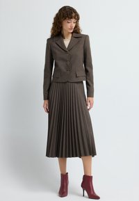 Dopasowana brązowa marynarka z trzema guzikami, połączona z plisowaną brązową spódnicą i bordowymi botkami na obcasie, o gładkiej teksturze.