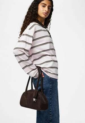 Giovane donna con capelli ricci che indossa una maglietta a maniche lunghe a righe e jeans blu, con una borsa in pelle marrone scuro tenuta al fianco.