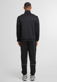 Mann steht mit dem Rücken zu sehen, trägt einen schwarzen Trainingsanzug mit weißer Trim an der Hose und grauen Laufschuhen mit weißen Socken.