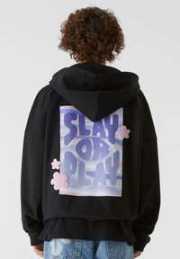 Svart hettegenser med et stort ryggramme som leser "SLAY OR PLAY" i lilla og rosa, med sky-lignende mønstre og rosa detaljer.