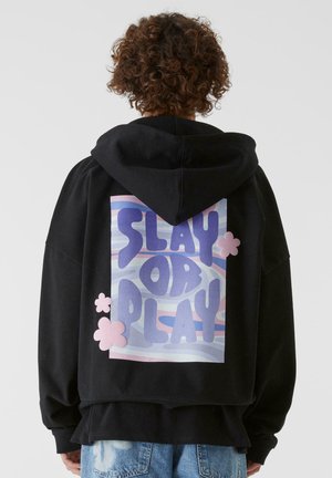 Sort hættebluse med et stort rygtryk, der står "SLAY OR PLAY" i lilla og lyserød, med skylignende mønstre og lyserøde accenter.