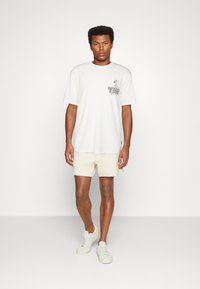 T-shirt blanc à manches courtes avec imprimé graphique noir sur la poitrine, associé à un short texturé crème et des baskets de couleur claire.