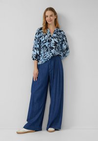 Donna in piedi contro un muro bianco che indossa una blusa floreale blu, pantaloni in denim a gamba larga e sneakers bianche, sorridendo dolcemente.