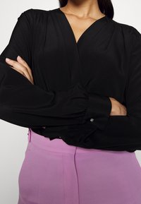 Blusa nera avvolgente con maniche lunghe e ampie, caratterizzata da un scollo a V e dettagli arricciati sulle spalle. Abbinata a pantaloni rosa chiaro.