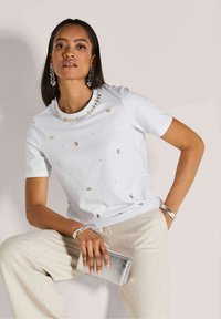 Witte t-shirt met korte mouwen en versierde halslijn met kleine gouden accenten, gecombineerd met lichtbeige broeken en een zilveren clutch.
